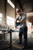 A person wearing safety equipment drills into a metal beam in a workshop.