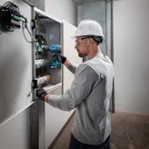 A person wearing safety equipment uses a cordless drill on an electrical panel.