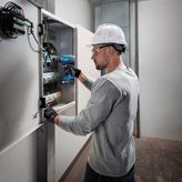 A person wearing safety equipment uses a cordless drill on an electrical panel.