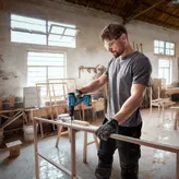 A person wearing safety equipment drills into a wooden frame in a workshop.