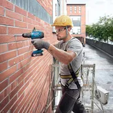 A person wearing safety equipment drills into a brick wall on scaffolding.