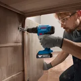 A person wearing safety equipment uses a cordless drill to install a cabinet hinge.