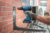 A person wearing safety equipment uses a cordless drill to fasten a metal bracket to a brick wall.
