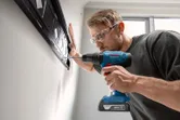 A person wearing safety equipment drills into a wall to install a mounting bracket.