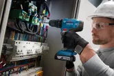 A person wearing safety equipment drills into a control panel full of wires.