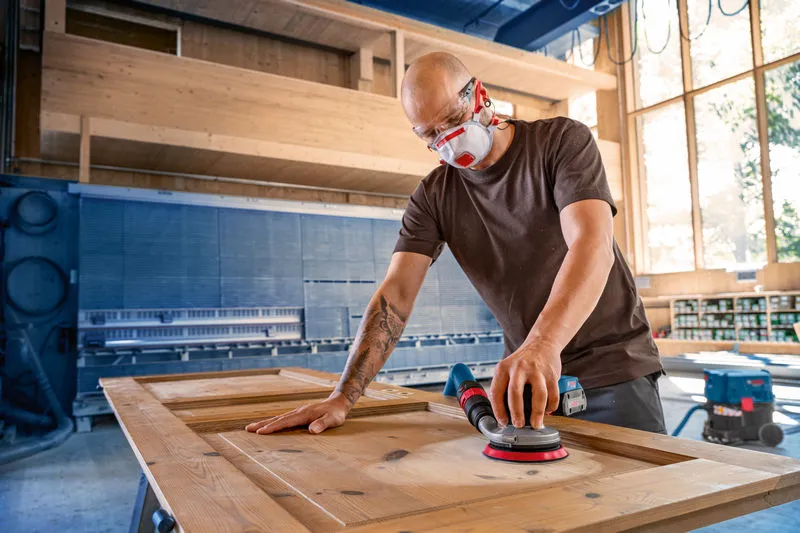 A person wearing safety equipment sands a wooden door with a cordless random orbit sander.