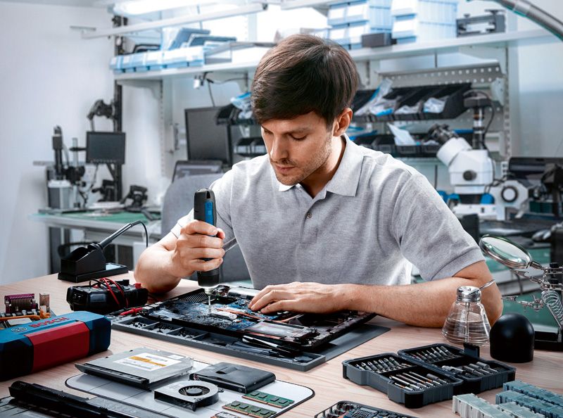 A person uses a cordless screwdriver to assemble electronic components on a workbench.