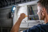 A person uses a screwdriver on an electrical panel, cordless light attached for illumination.