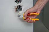 A person uses insulated pliers to cut a green wire from an electrical wall box.