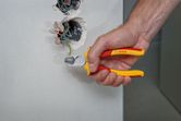 A person uses insulated pliers to cut a green wire from an electrical wall box.