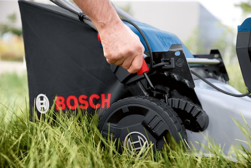 Person adjusts the height lever on a cordless lawn mower set in tall grass.