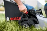 Person adjusts the height lever on a cordless lawn mower set in tall grass.