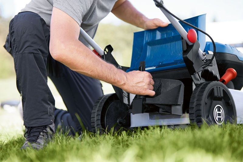 A person adjusts the compartment of a cordless lawn mower on a grassy lawn.
