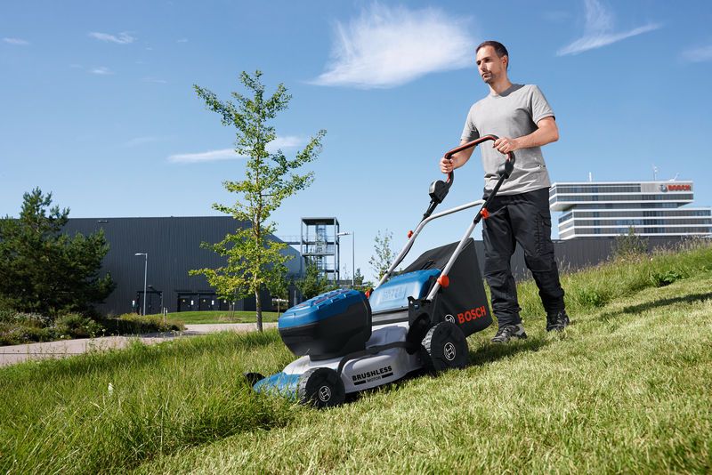 A person pushes a cordless lawn mower across a grassy slope outdoors.