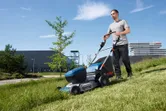 A person pushes a cordless lawn mower across a grassy slope outdoors.