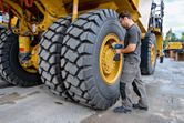 A person wearing safety equipment uses an impact wrench on a large construction vehicle tire.