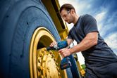 A person wearing safety equipment uses an impact wrench on a large vehicle wheel.