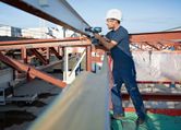 A person wearing safety equipment uses a cordless impact wrench on a steel beam.