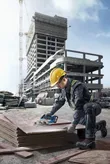 A person wearing safety equipment cuts plywood on a construction site with a cordless saw.