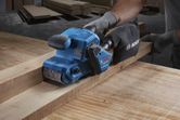 A person wearing safety equipment sands a wooden plank with a blue belt sander.