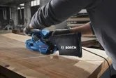 A person wearing safety equipment sands a wooden board with a blue belt sander.