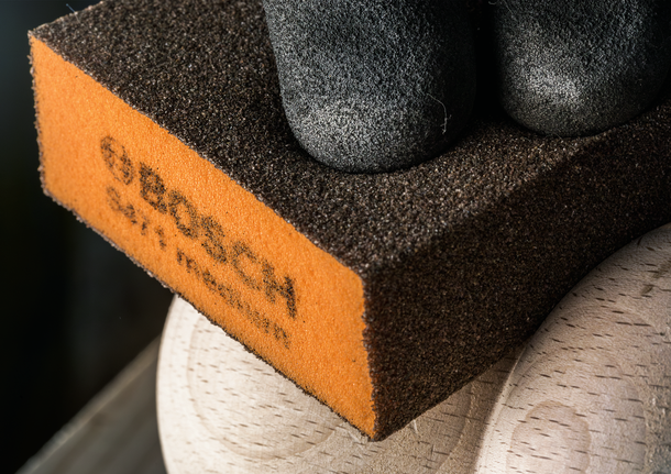 Hand in a black glove holds a sanding sponge against a wooden surface.