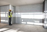 A person wearing safety equipment uses a laser leveling tool to project a straight line on a wall.
