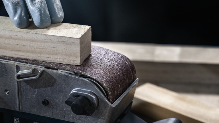 Person wearing safety equipment sanding a wooden block with a belt sander.