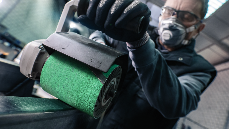 Person wearing safety equipment polishes a metal surface with a power sander.