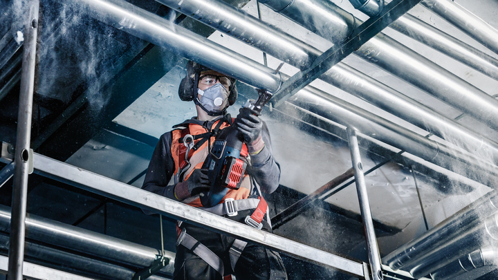 Object #200456602: 3046762_app_web_1 Person wearing safety equipment drills into a metal pipe on an overhead structure.