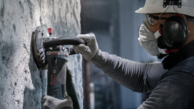 A person wearing safety equipment uses a power grinder to sand a concrete wall.