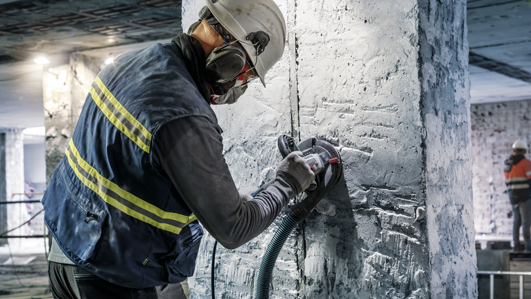Person wearing safety equipment operates a power tool to grind concrete on a construction site.