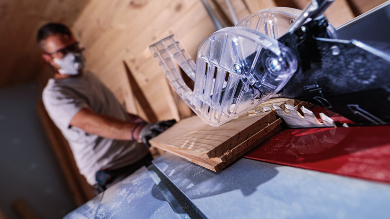 A person wearing safety equipment cuts wood with a table saw.