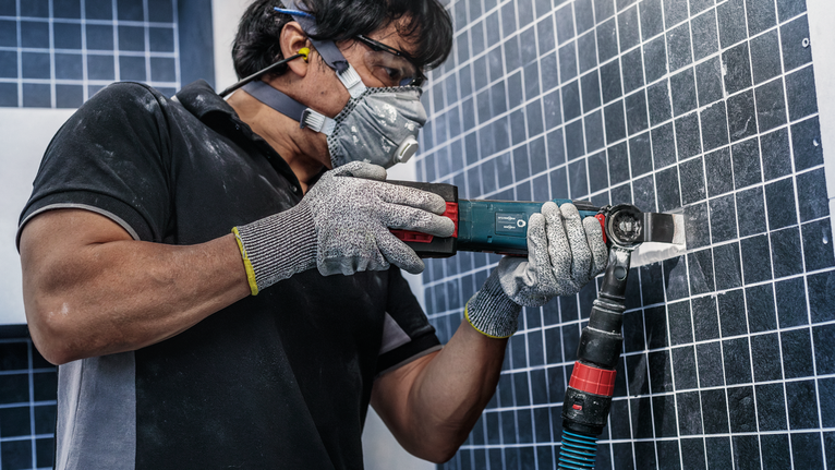 A person wearing safety equipment uses a power tool to cut tiles on a wall.