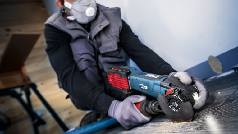 PRO ACZ 85 EIB Multitool Blade, 85 mm Person wearing safety equipment uses an angle grinder to cut flooring near a wall.