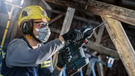 A person wearing safety equipment uses a power drill on a wooden beam.