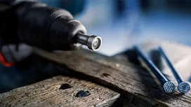 Power tool driving a fastener into wood next to several large nails.
