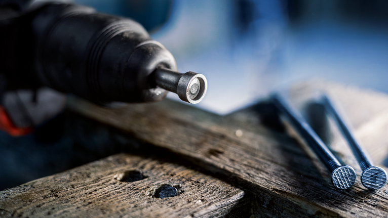 Power tool driving a fastener into wood next to several large nails.