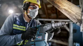 A person wearing safety equipment drills into a wooden beam with a power tool.