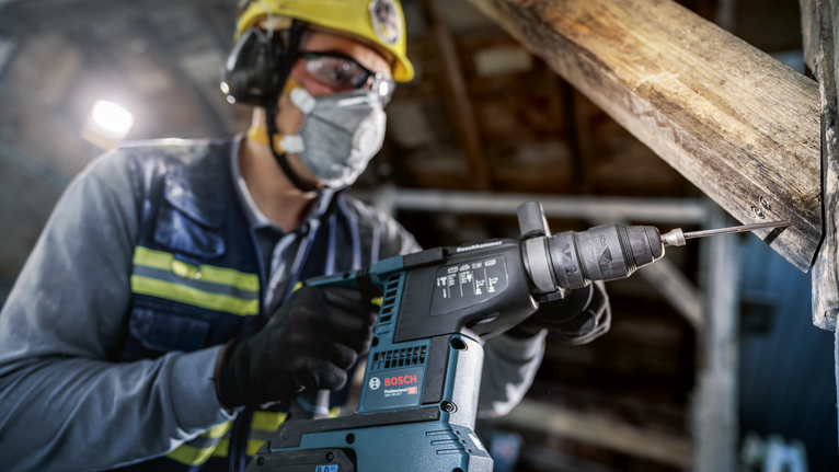 A person wearing safety equipment drills into a wooden beam with a power tool.