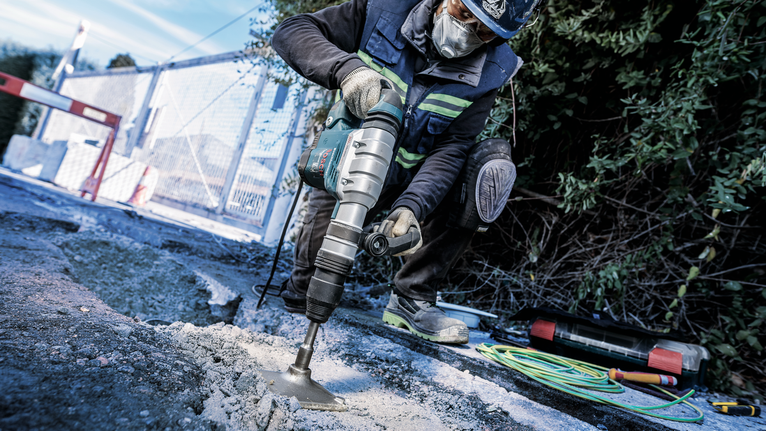 PRO SDS max Tamping Plate, 150 x 150 mm A person wearing safety equipment drills into pavement at a construction site.