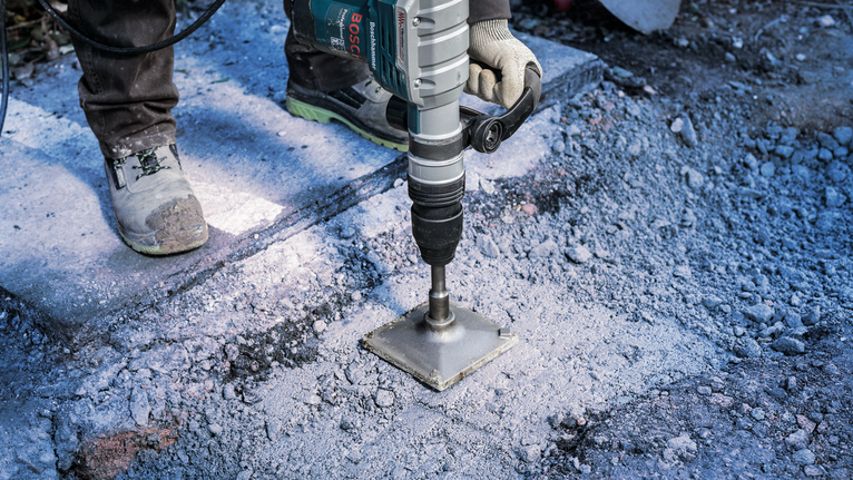 PRO SDS max Tamping Plate, 150 x 150 mm Person wearing safety equipment uses a power drill to break concrete outdoors.