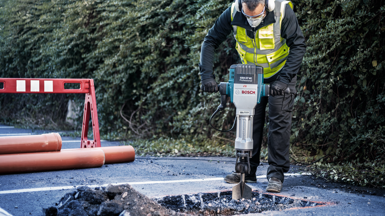 A person wearing safety equipment breaks asphalt on a road with a demolition hammer.