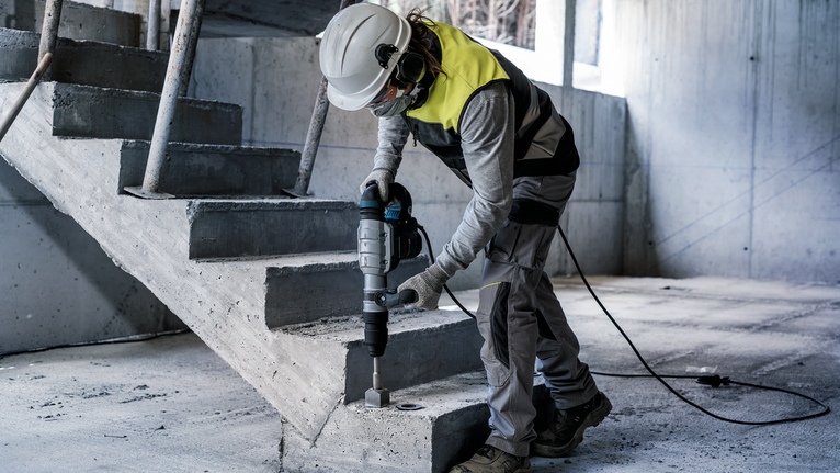 Person wearing safety equipment drills into concrete near unfinished stairs.