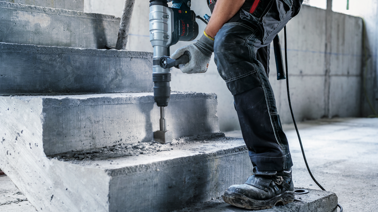Person wearing safety equipment uses a power tool to break concrete stairs.