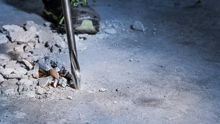 Rotary hammer drilling into concrete floor near scattered debris.