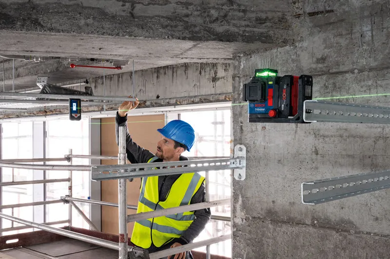 A person wearing safety equipment aligns metal framing using a laser leveling tool with a laser receiver.