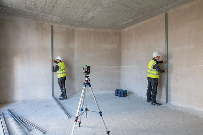 Two workers wearing safety equipment align metal studs using a laser leveling tool on a tripod.