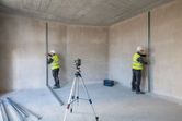 Two workers wearing safety equipment align metal studs using a laser leveling tool on a tripod.