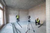 Two people wearing safety equipment align metal studs using a laser leveling tool in an unfinished room.